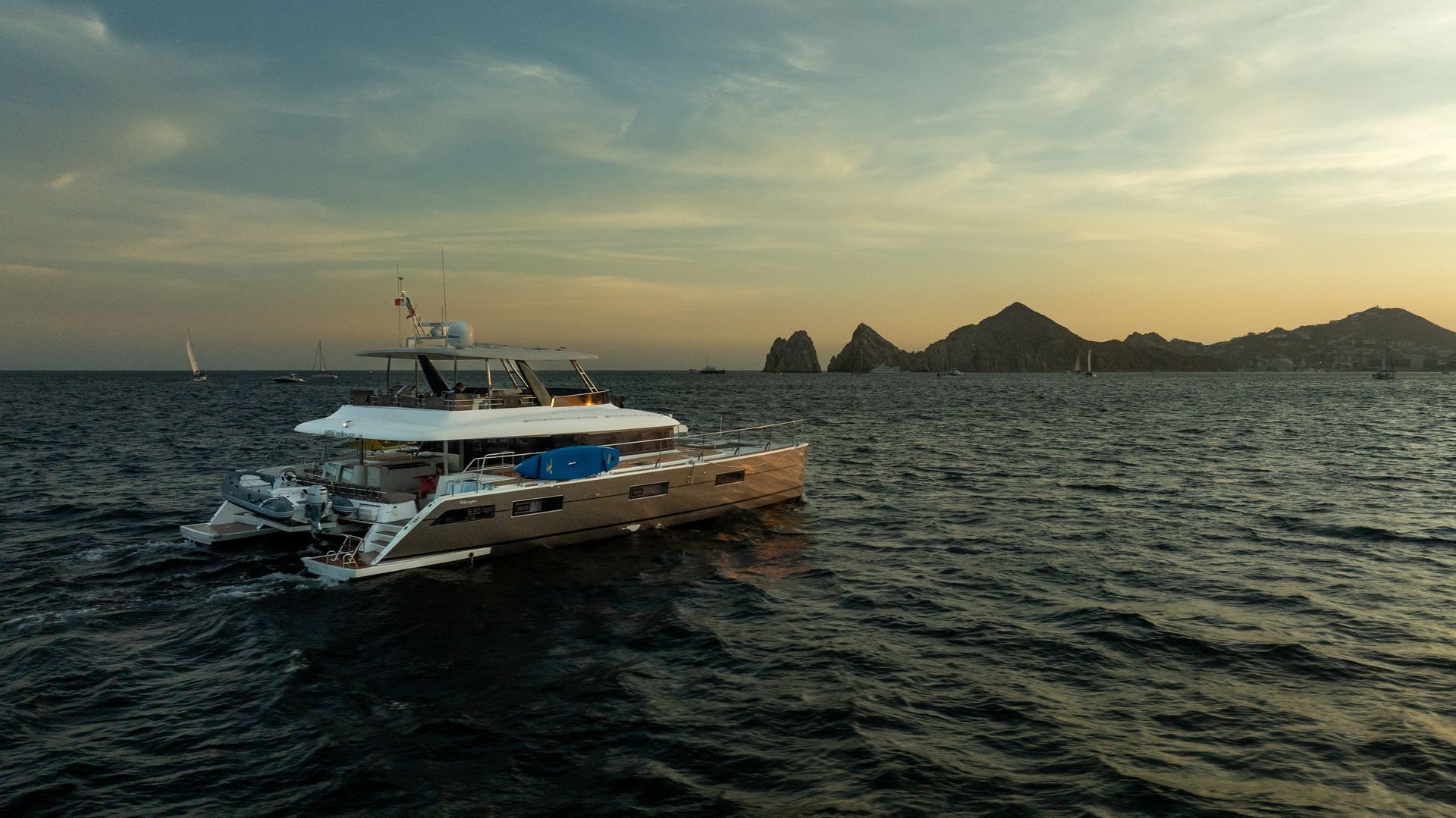 Catamaran sailing on the ocean with rocky hills in the background during sunset.
