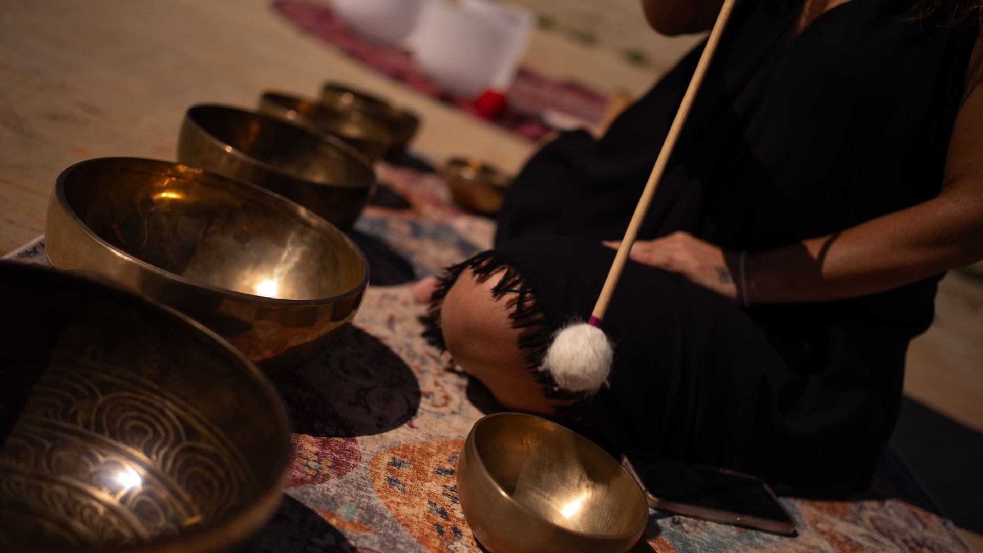 Person playing singing bowls with a mallet on a patterned mat outdoors.