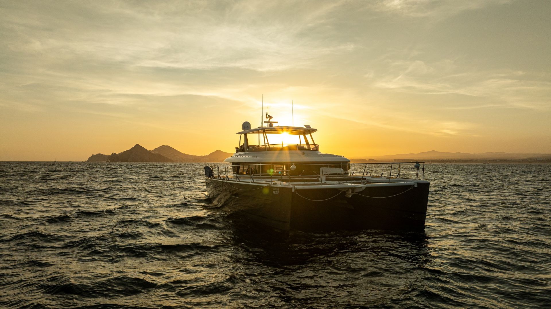 Catamaran boat sailing at sunset with mountains in the background and gentle waves on the water.